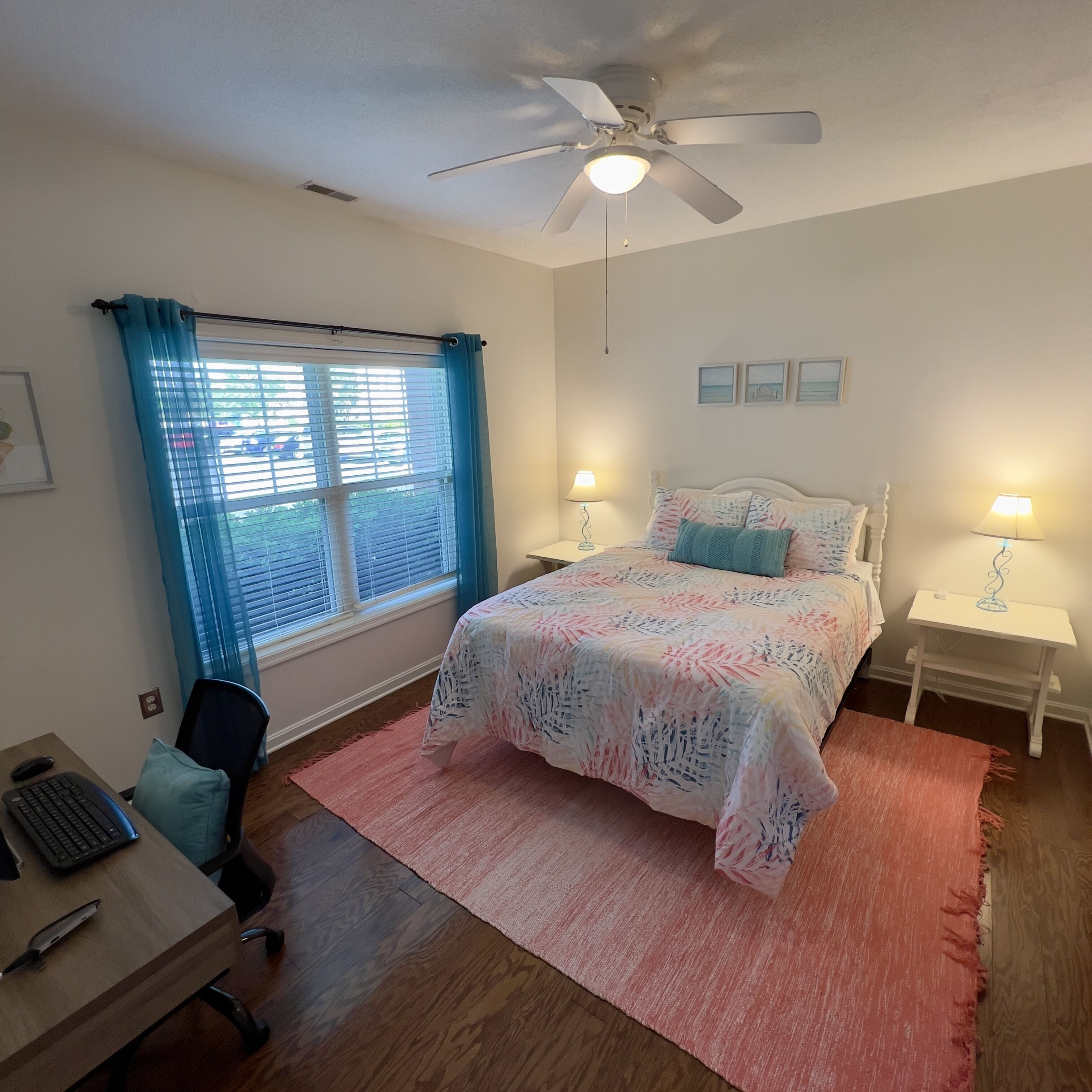 A bedroom with a bed, two bedside tables with lamps, a desk with a computer, a chair, a ceiling fan, and a window with curtains.