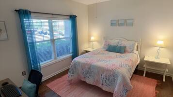 A bedroom with a bed, two bedside tables with lamps, a desk with a computer, a chair, a ceiling fan, and a window with curtains.