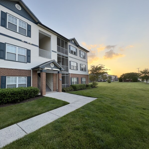 A multi-story residential building with a covered entrance, a grassy lawn, and a clear sky.