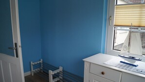 Ein Zimmer mit blauer Wand, einer weißen Tür, einer weißen Kommode mit Schubladen und einem Fenster mit Jalousien. Auf der Kommode steht ein kleines Modell eines Segelbootes.