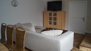 Ein Wohnzimmer mit einer Couch, zwei Korbstühlen, einem Fernseher auf einem Schrank und einer Tür.