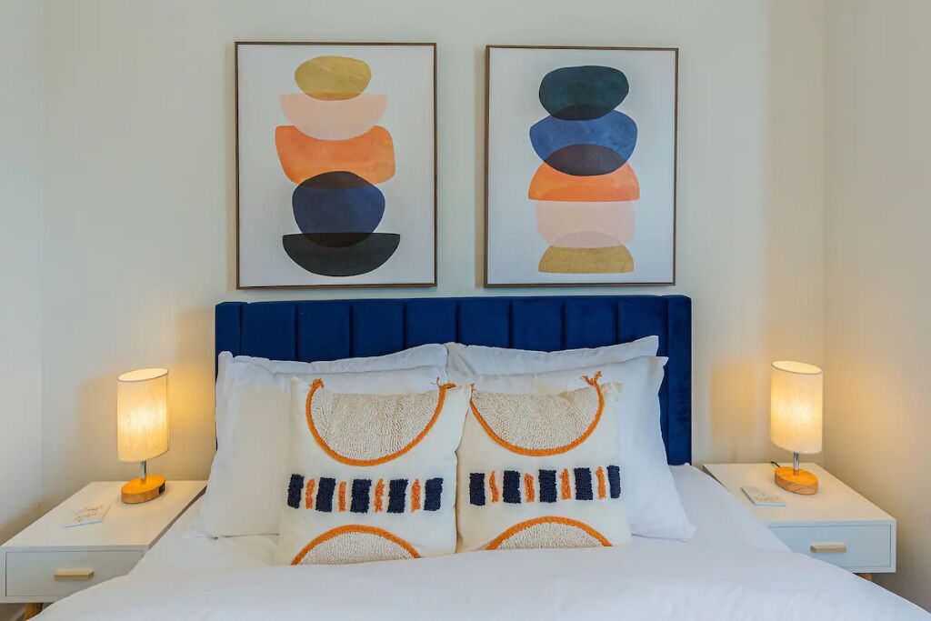 A hotel room with a bed, two bedside tables with lamps, a blue headboard, and two framed abstract paintings above the bed.