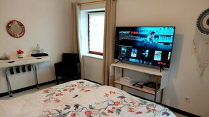 A bedroom with a bed, a desk, a chair, a TV, and a window.