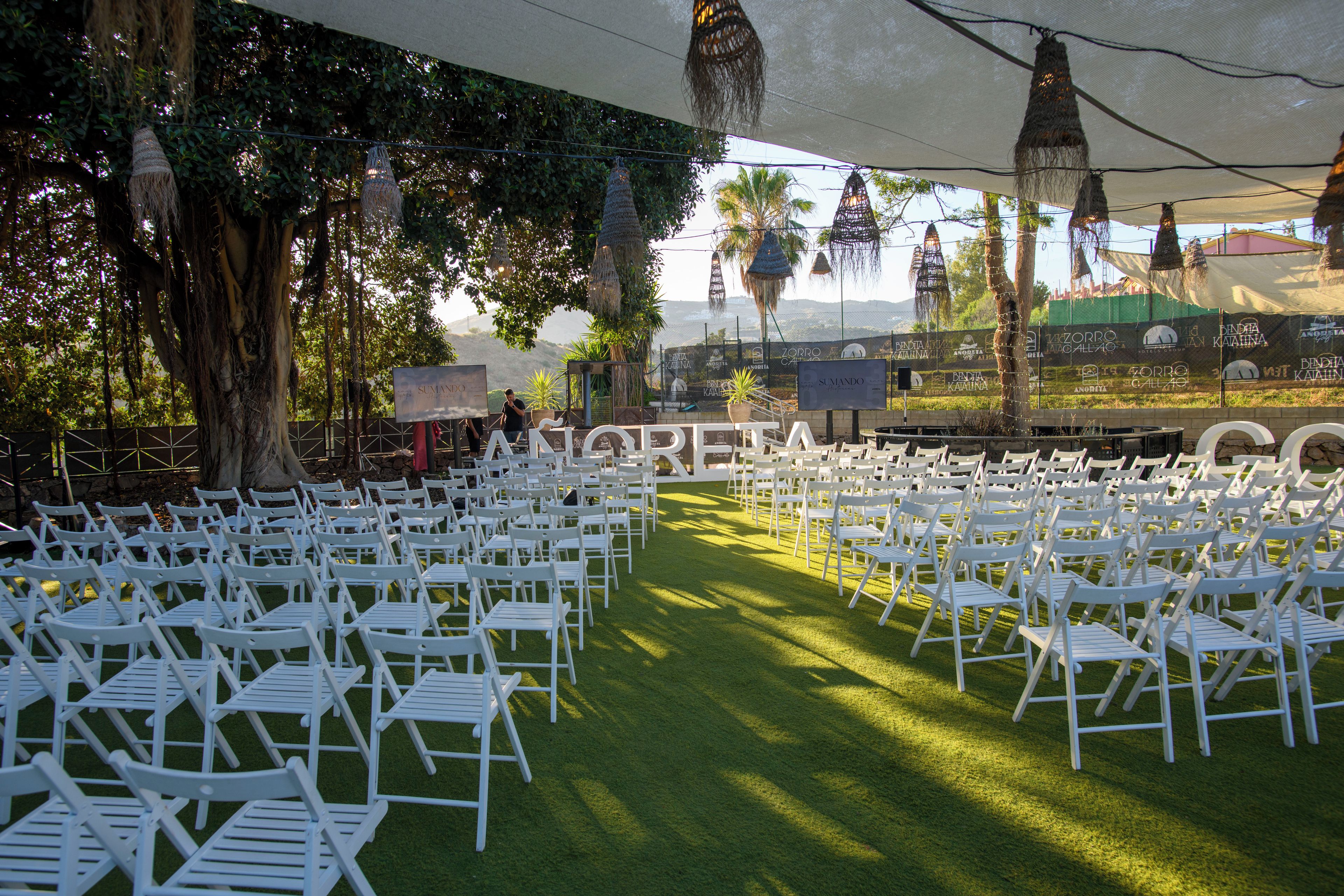 Un espacio para eventos al aire libre con filas de sillas blancas dispuestas sobre césped artificial, bajo un toldo grande con decoraciones colgantes.