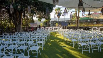 Un espacio para eventos al aire libre con filas de sillas blancas dispuestas sobre césped artificial, bajo un toldo grande con decoraciones colgantes.