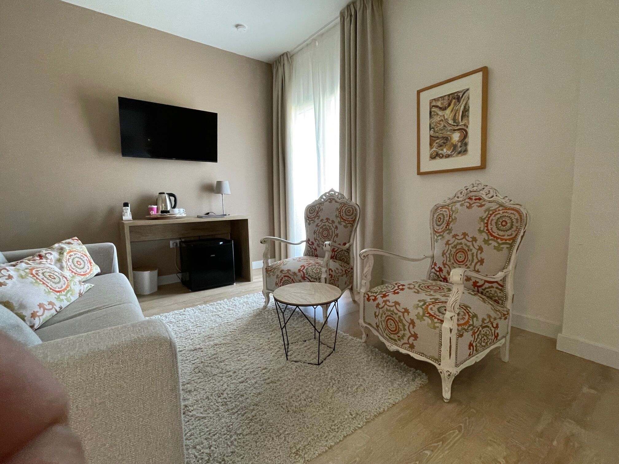 A living room with a sofa, two armchairs, a coffee table, a flat-screen TV, a desk with a lamp, and a painting on the wall.