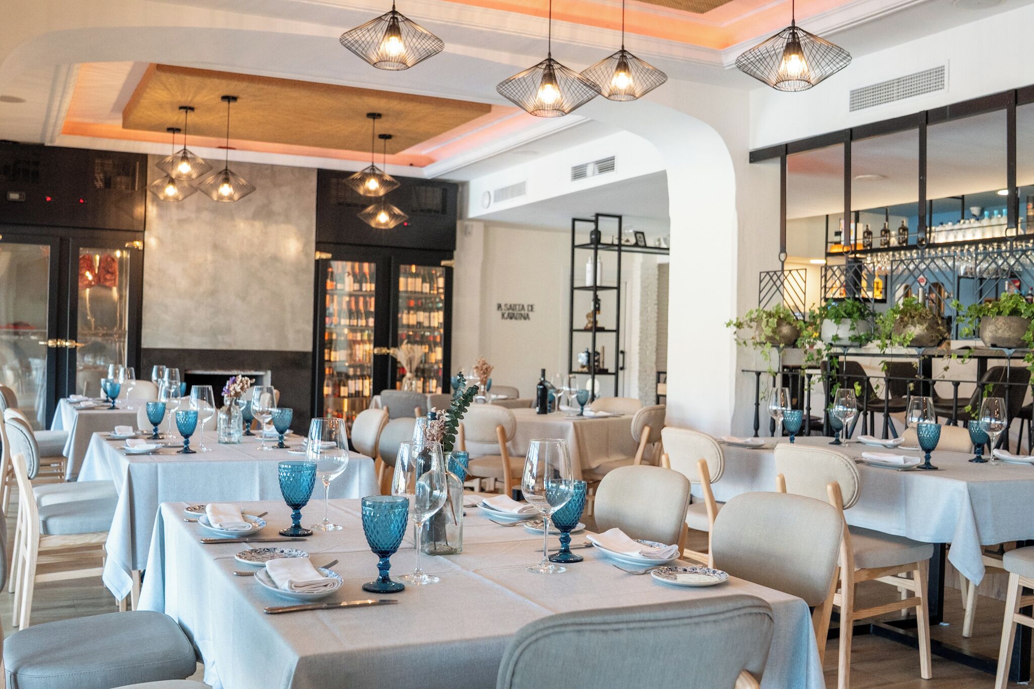 Un restaurante con mesas dispuestas para comer, lámparas colgantes y una zona de bar con botellas.