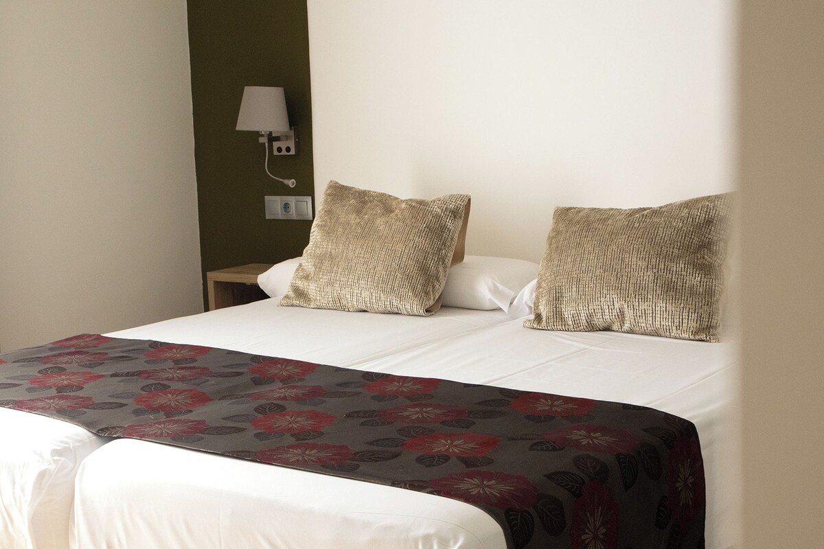 A bed with a floral bedspread, two textured pillows, a nightstand, a wall-mounted lamp, and a window with curtains.