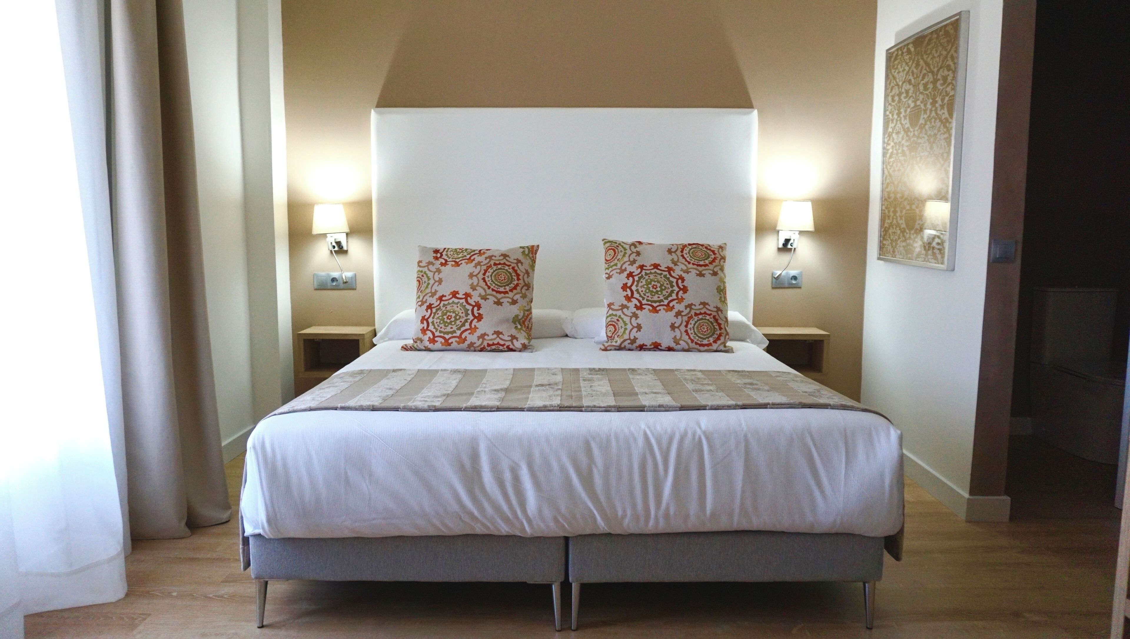 Una cama bien hecha con almohadas estampadas, dos mesitas de noche con lámparas y una obra de arte colgada en la pared.