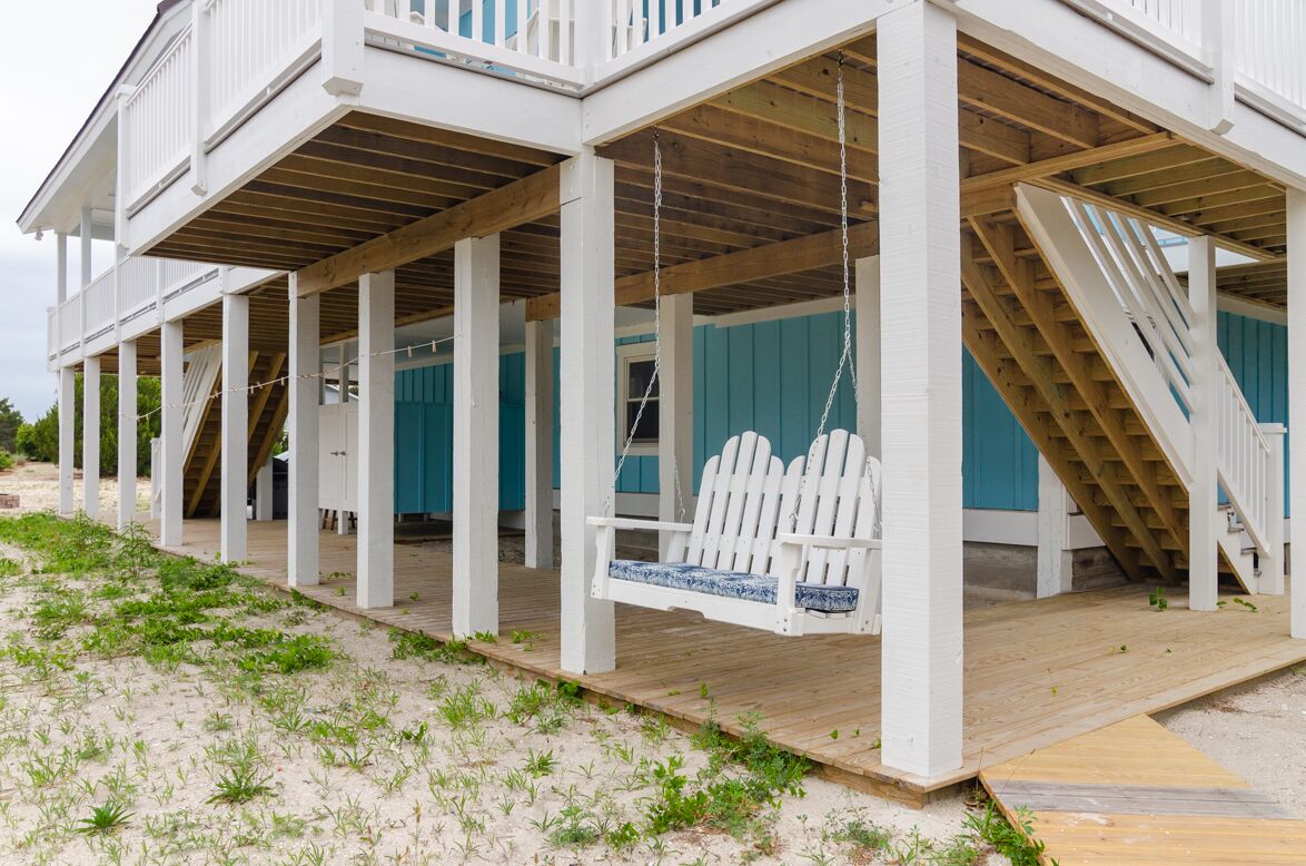 Ein Strandhaus mit einer Holzterrasse, einer Schaukel und einer Treppe, die zu einer oberen Ebene führt.