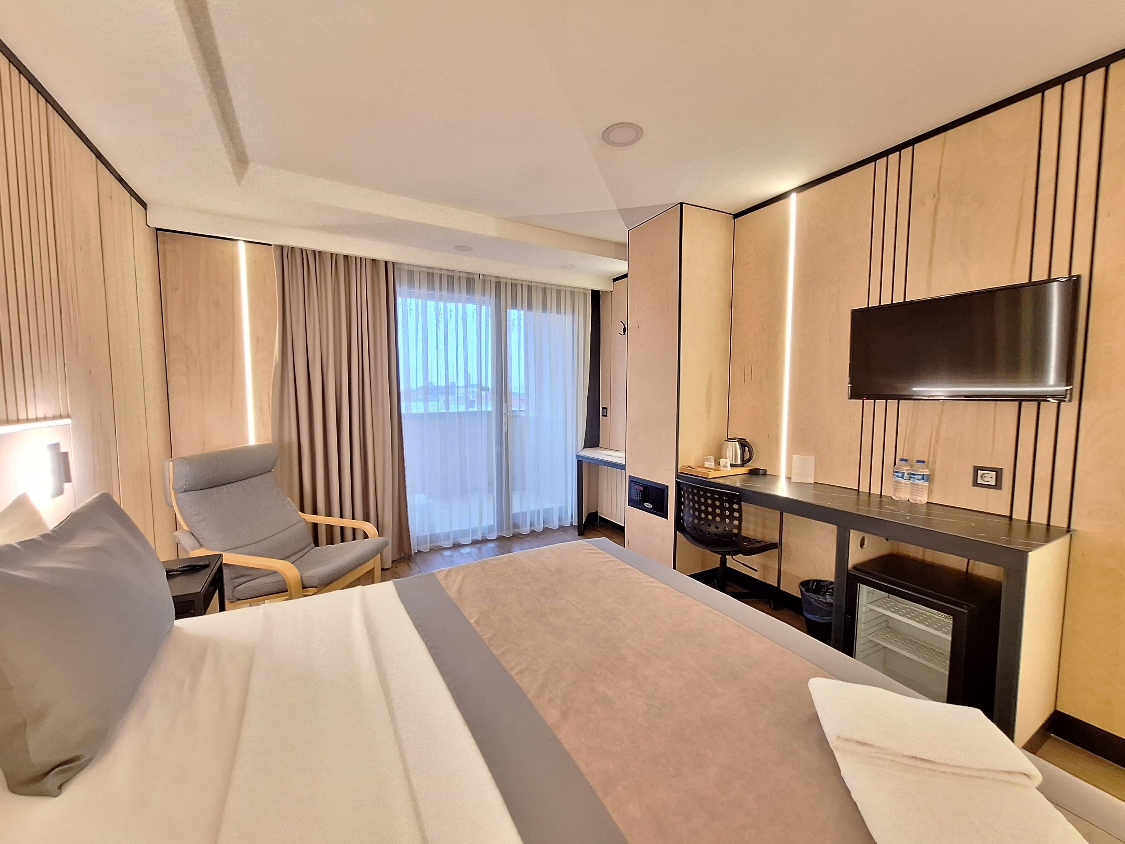 A modern hotel room with a large bed, a desk, a chair, a TV, and a view of the outdoors.