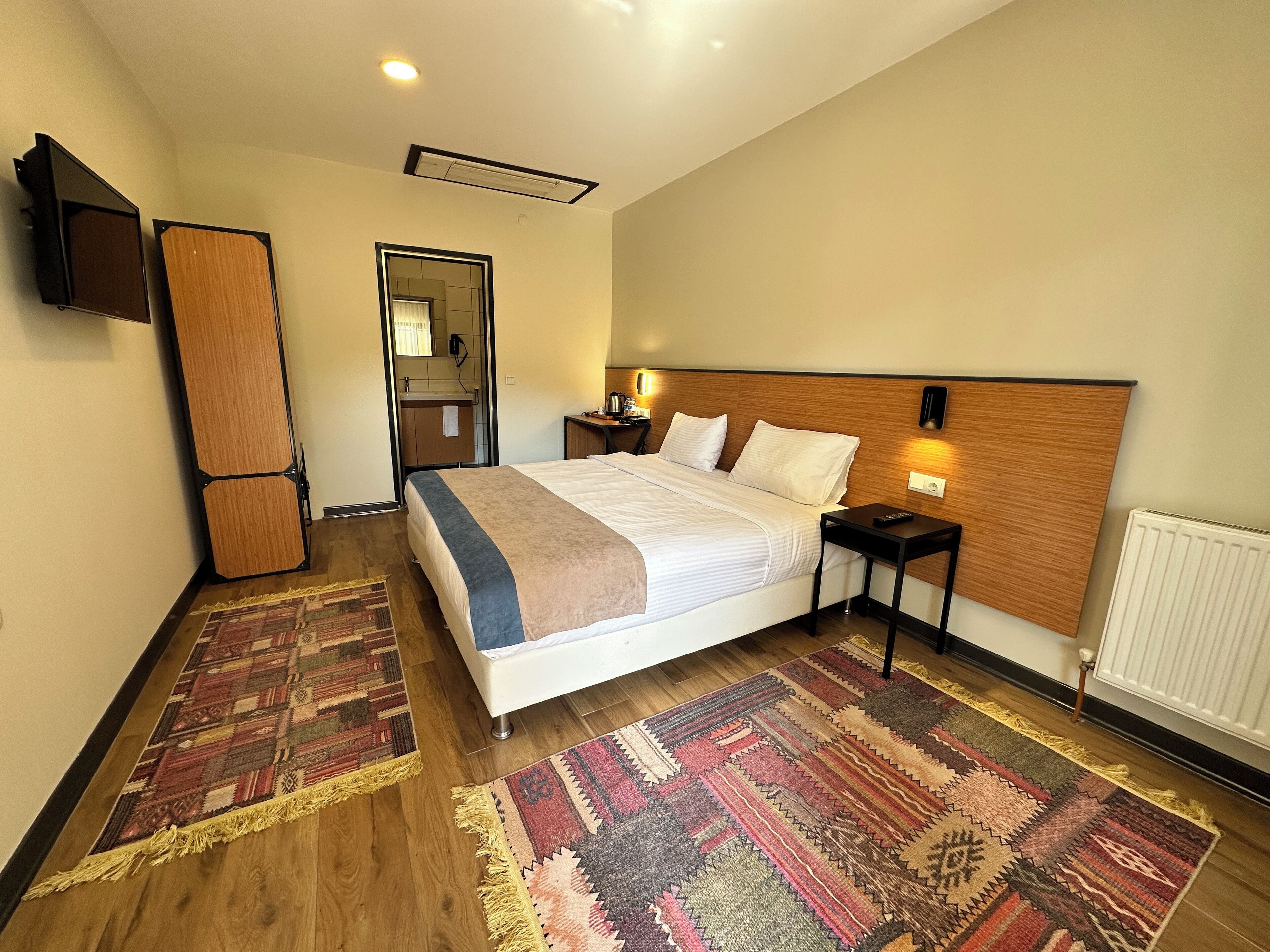 Habitación de hotel con una cama grande, una mesita de noche con lámpara, un televisor montado en la pared y un cabecero de madera.