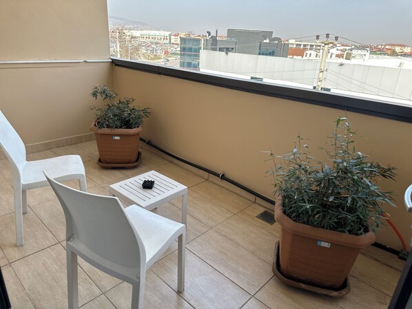 Un balcón con dos sillas blancas, una mesita pequeña y dos plantas en macetas.