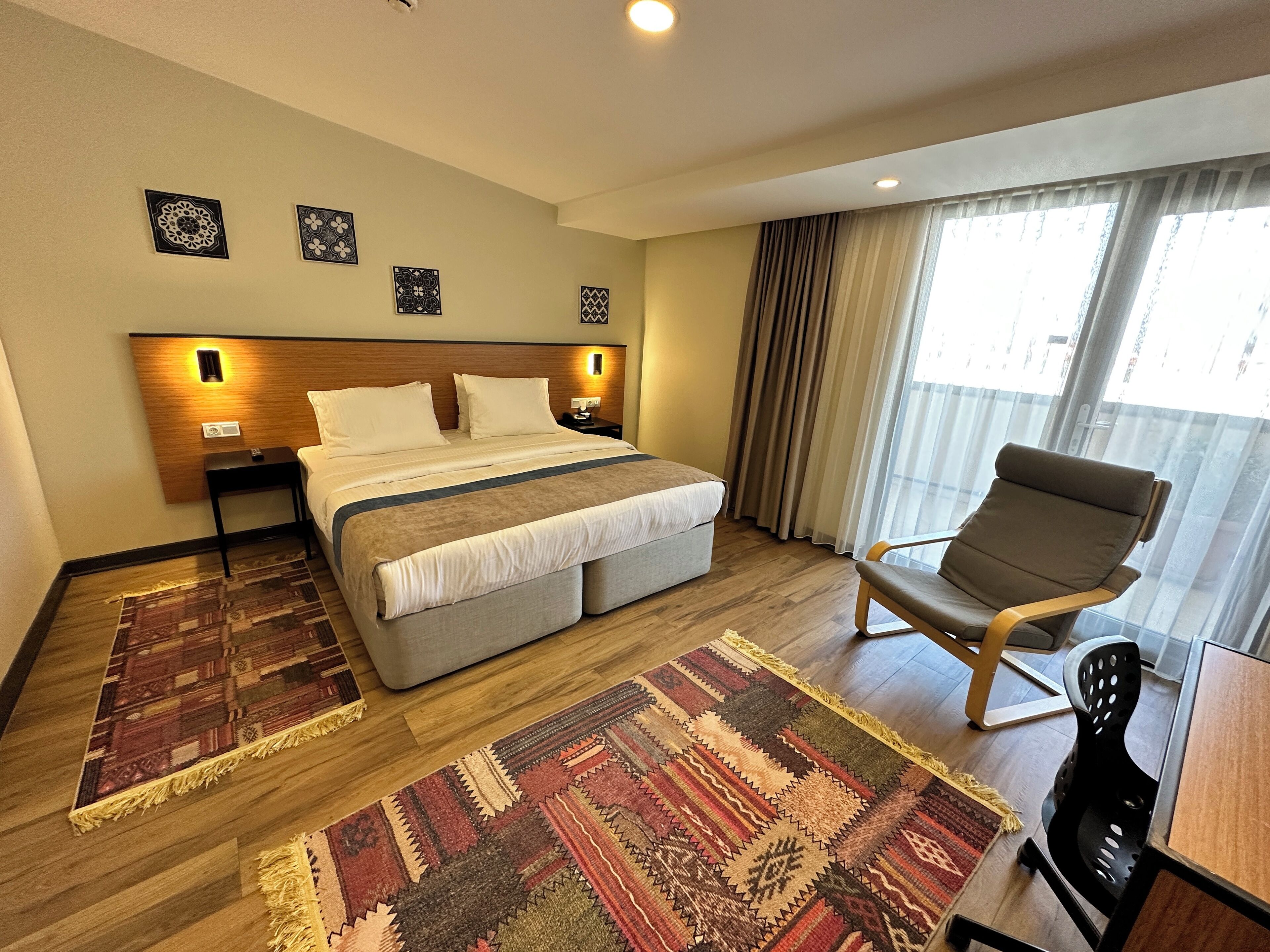 A hotel room with a large bed, a chair, a small table, a nightstand, and a window with curtains.