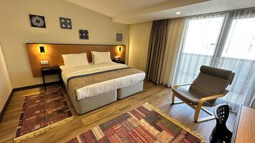 A hotel room with a large bed, a chair, a small table, a nightstand, and a window with curtains.