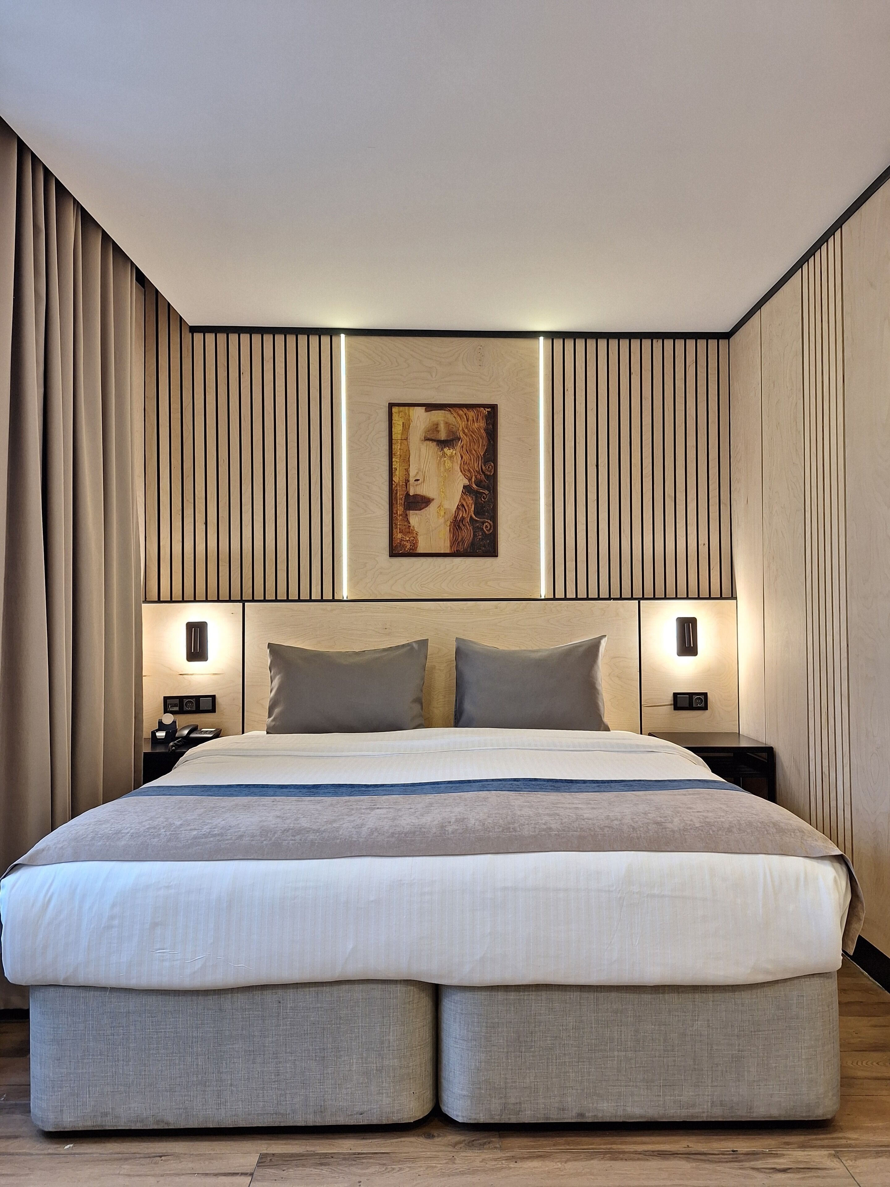 A modern hotel room with a large bed, two bedside tables, a wall-mounted artwork, and a curtain-covered window.