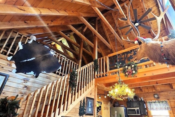 Interior de una cabaña de madera con techo alto, vigas de madera, una escalera y un candelabro.