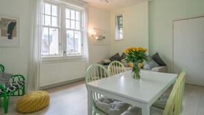 Une pièce lumineuse avec une table blanche, des chaises vertes, un pouf jaune et un vase rempli de fleurs jaunes.