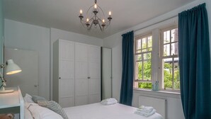 Une chambre à coucher avec un lit, une table de chevet, une lampe, un ours en peluche, une armoire et une fenêtre avec des rideaux.