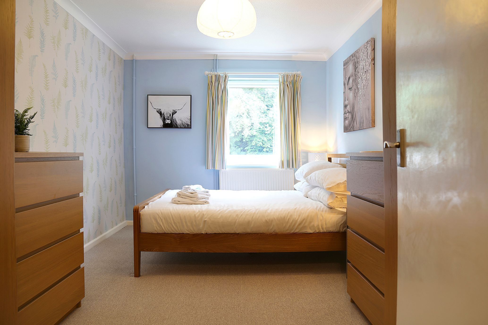 Un dormitorio ordenado con cama, cómoda, ventana con cortinas y una obra de arte colgada en la pared.