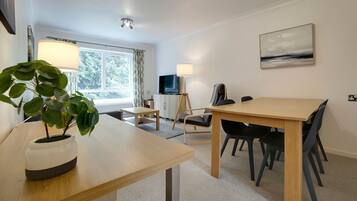A modern room with a wooden table, black chairs, a potted plant, a standing lamp, a TV, and a painting on the wall.