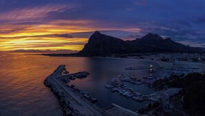 A coastal marina at sunset with boats docked, a breakwater, and mountains in the background.