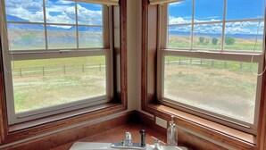 Un bany amb dues finestres grans que donen vista a un camp de gespa i muntanyes allunyades.