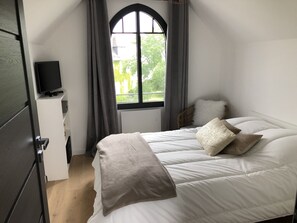 A bedroom with a bed, a television on a stand, a window with curtains, and a view of greenery outside.