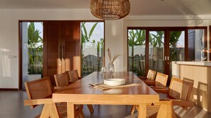 Een eethoek met een houten tafel en stoelen, een geweven lamp en grote schuifdeuren met uitzicht op groen.