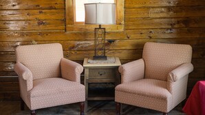 Deux fauteuils à motif chevrons, une petite table en bois avec une lampe et un tapis à carreaux sur un sol en pierre, dans une pièce d’une cabane en rondins.