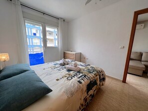 Ett sovrum med en säng, ett nattduksbord, en lampa och utsikt över byggnader genom fönstret.