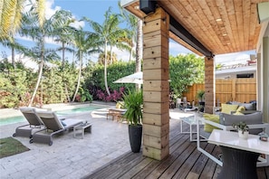 Una terraza de madera con piscina, sillas de descanso y un área de estar cubierta con cojines.