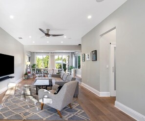 A spacious living room with a flat-screen TV, a ceiling fan, and a large window offering a view of the outdoors.
