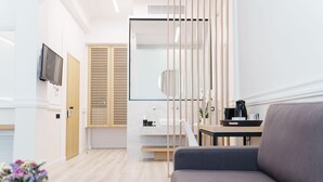 A modern hotel room with a sofa, a small table with a coffee maker, a TV mounted on the wall, and a wooden door. - Agora Athens Suites (Athens)