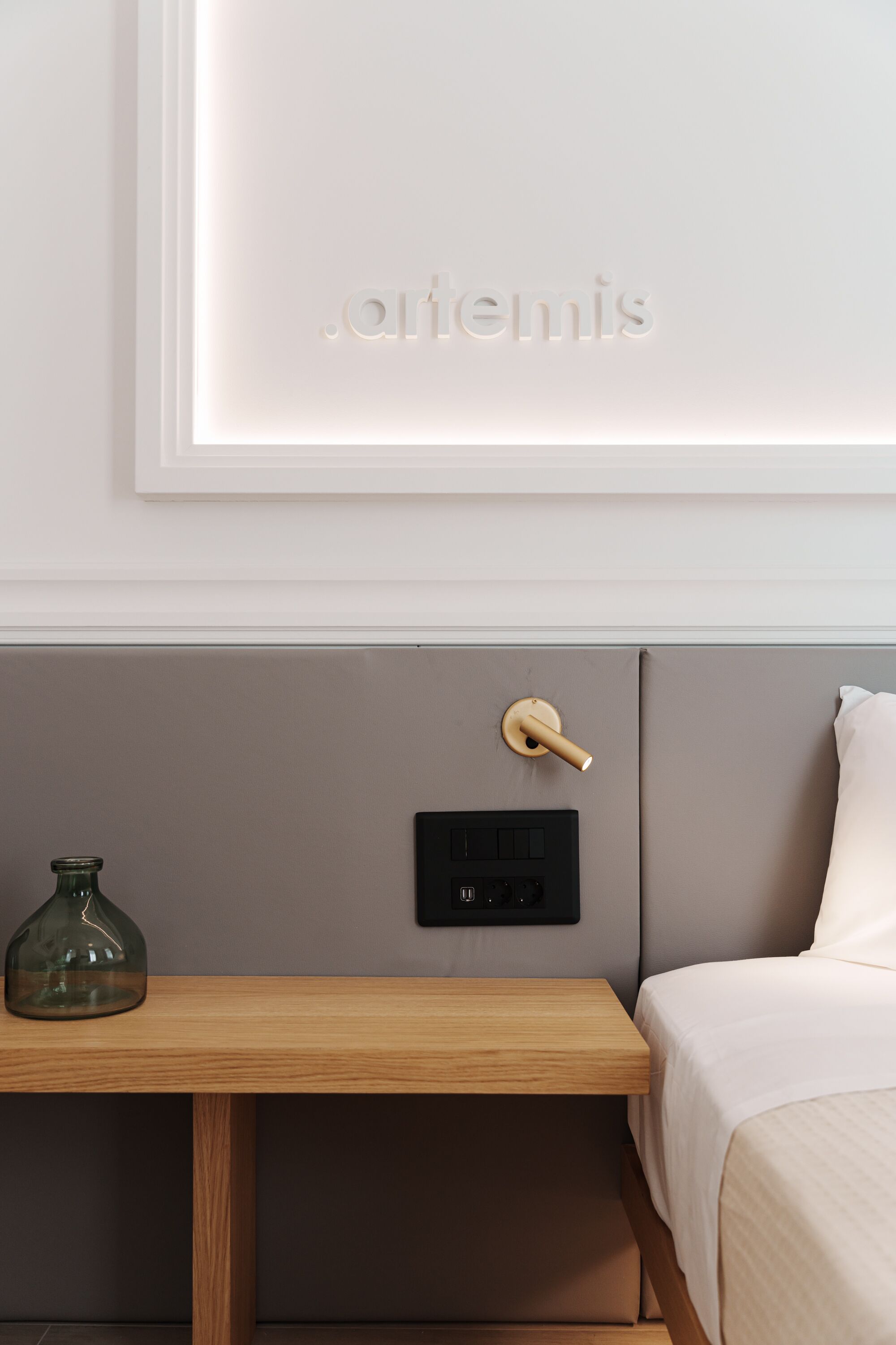 Een moderne hotelkamer met een minimalistisch design, voorzien van een nachtkastje, een in de muur gemonteerde lichtschakelaar en een bed met een hoofdeinde in neutrale tinten.