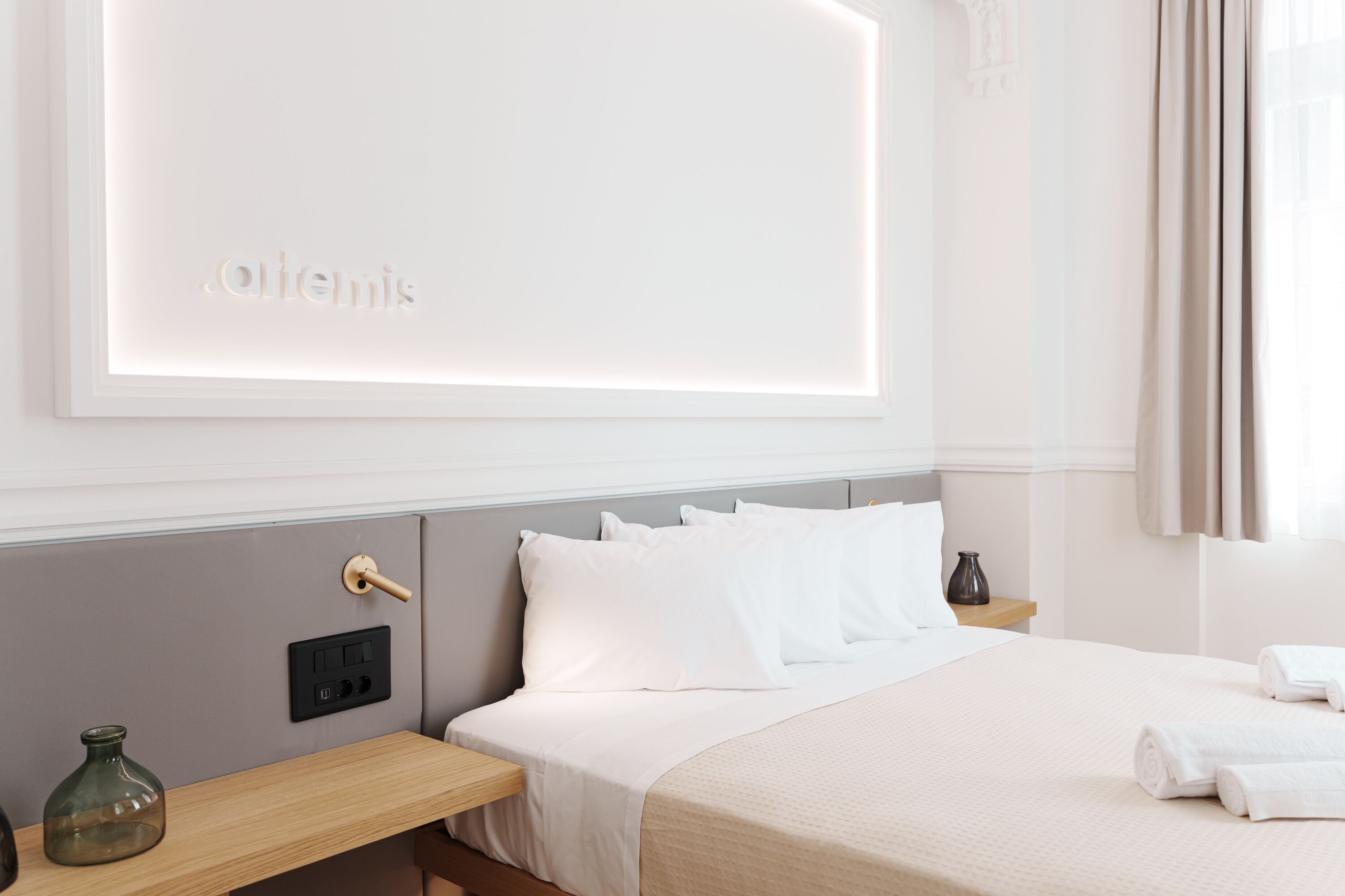 Una camera d'albergo con un letto, un comodino, una lampada a parete e una parete con pannello a incasso che mostra la scritta 'Athens'.