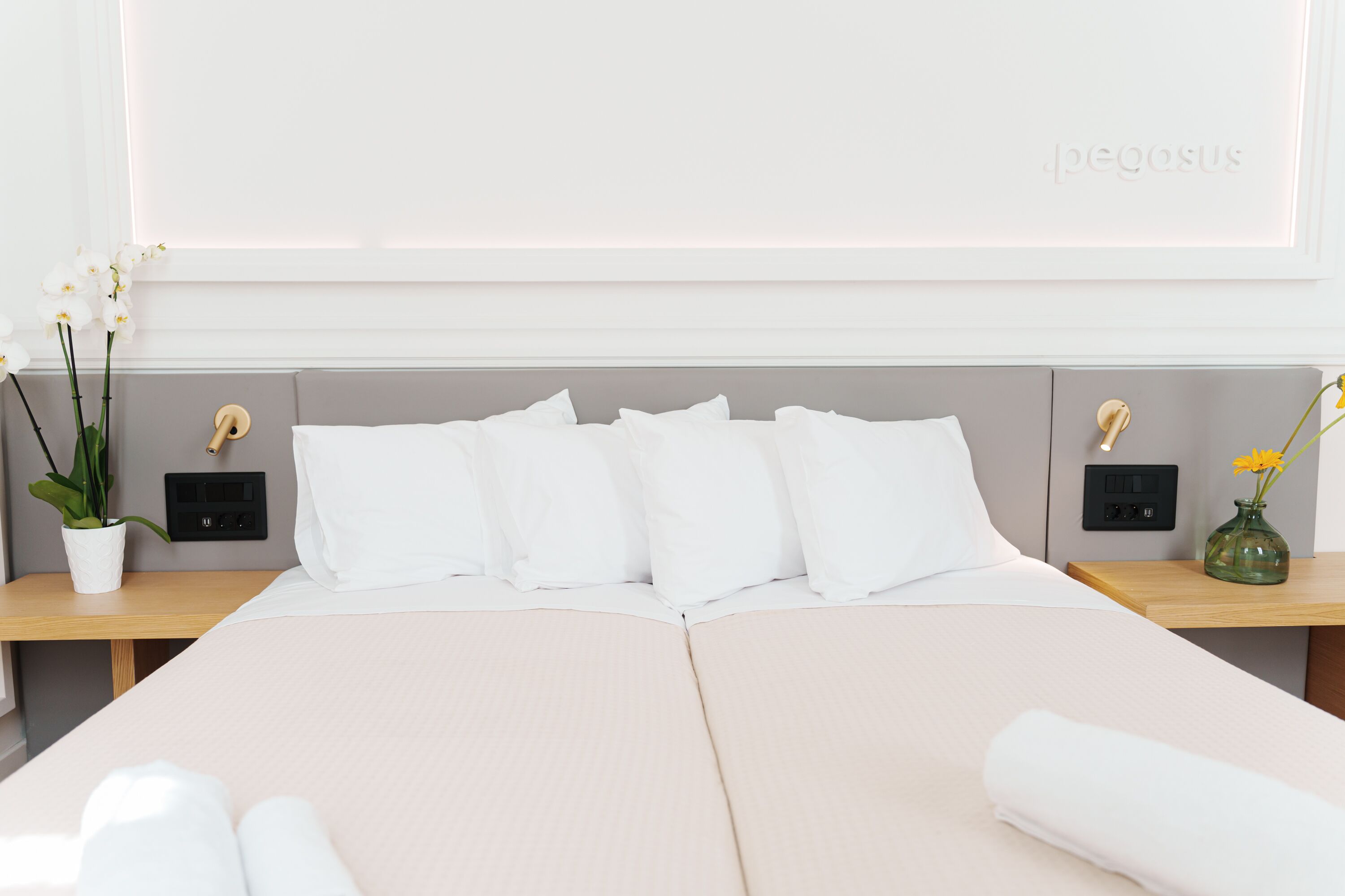 A neatly made bed with white linens, a bedside table with a vase of flowers, and a wall-mounted light switch.