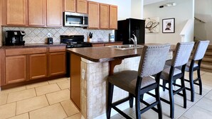 Una cocina con gabinetes de madera, un microondas, una estufa y un lavamanos. Hay cuatro taburetes de bar y una escalera que sube a otro nivel.