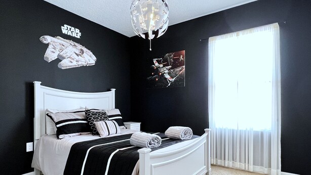 Un dormitorio con una cama blanca, ropa de cama blanco y negro, decoración de pared temática de Star Wars y un ventanal con cortinas translúcidas.
