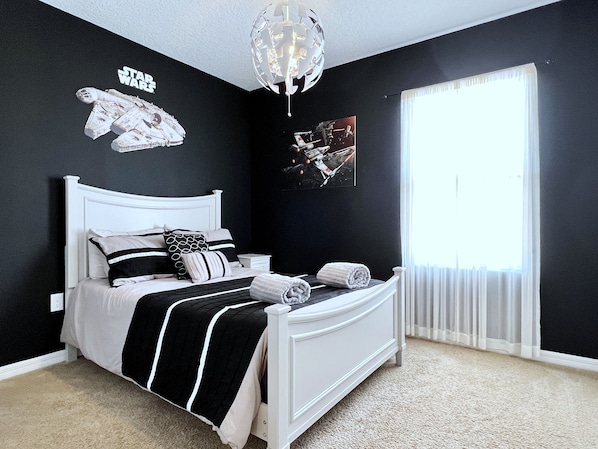 Ein Schlafzimmer mit einem weißen Bett, schwarz-weißer Bettwäsche, Wandbildern zum Thema Star Wars und einem großen Fenster mit transparenten Vorhängen.