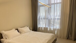 Siististi pedattu sänky valkoisilla lakanoiden kanssa huoneessa, jossa on suuri ikkuna, verhot ja puulattia.