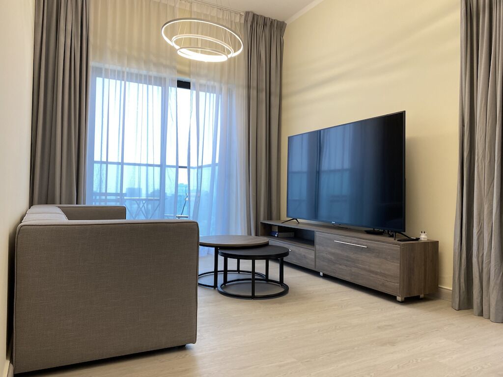 A modern living room with a sofa, a round table, a flat-screen TV, and a large window with curtains.