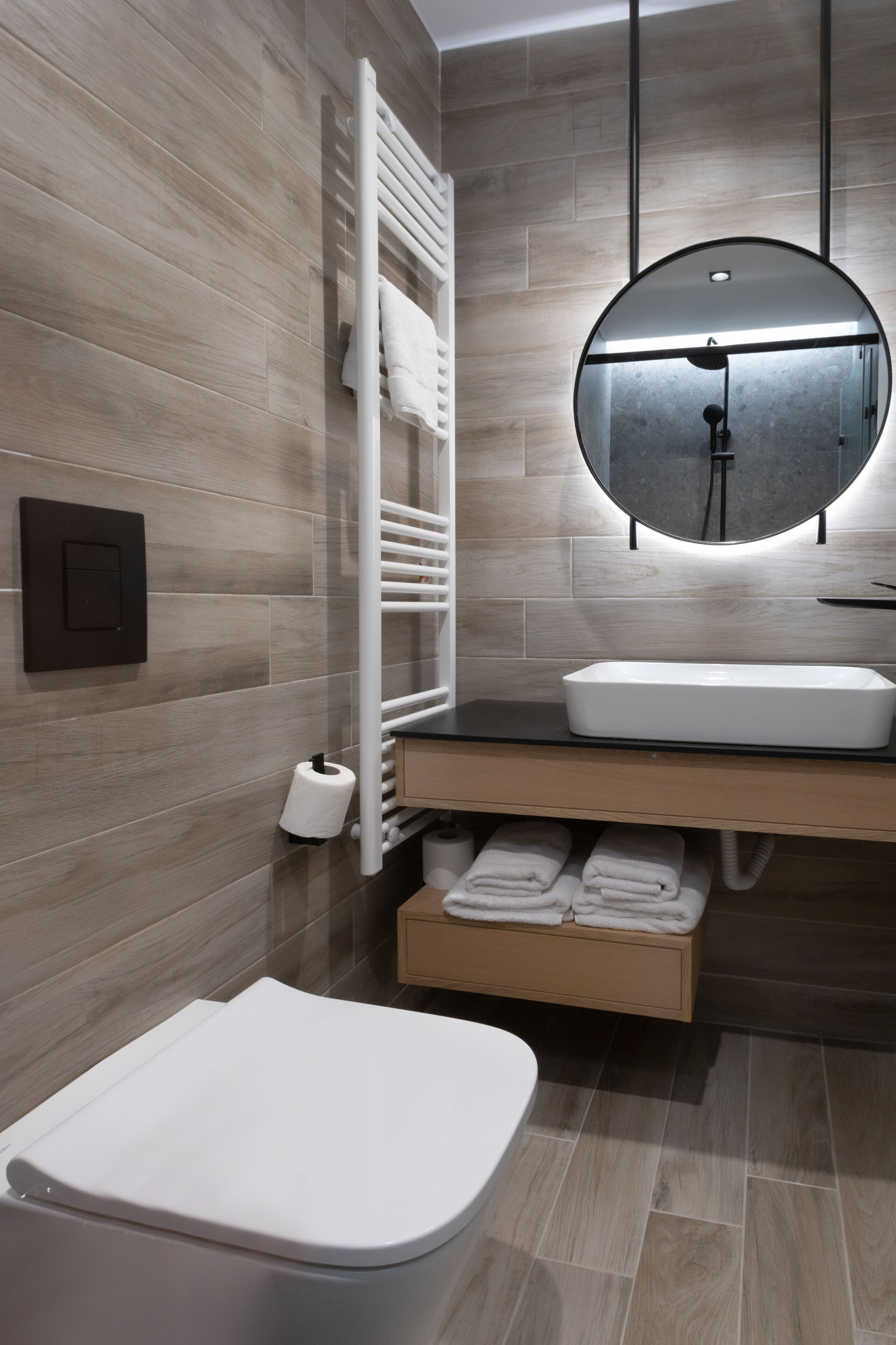 A modern bathroom with a wooden floor, a white toilet, a sink with a mirror above it, and a towel rack with towels.