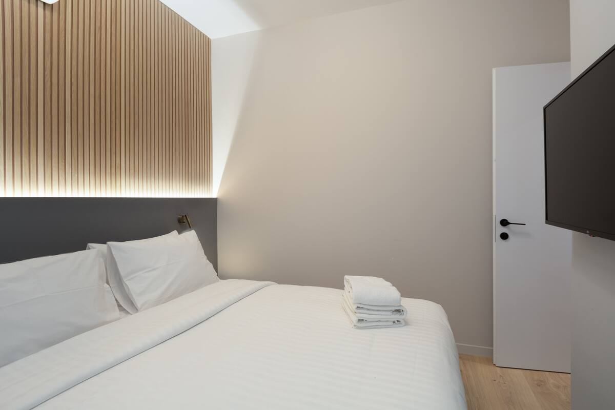 Una habitación de hotel moderna con una cama bien hecha, un cabecero de listones de madera, un televisor de pantalla plana montado en la pared y un armario blanco.