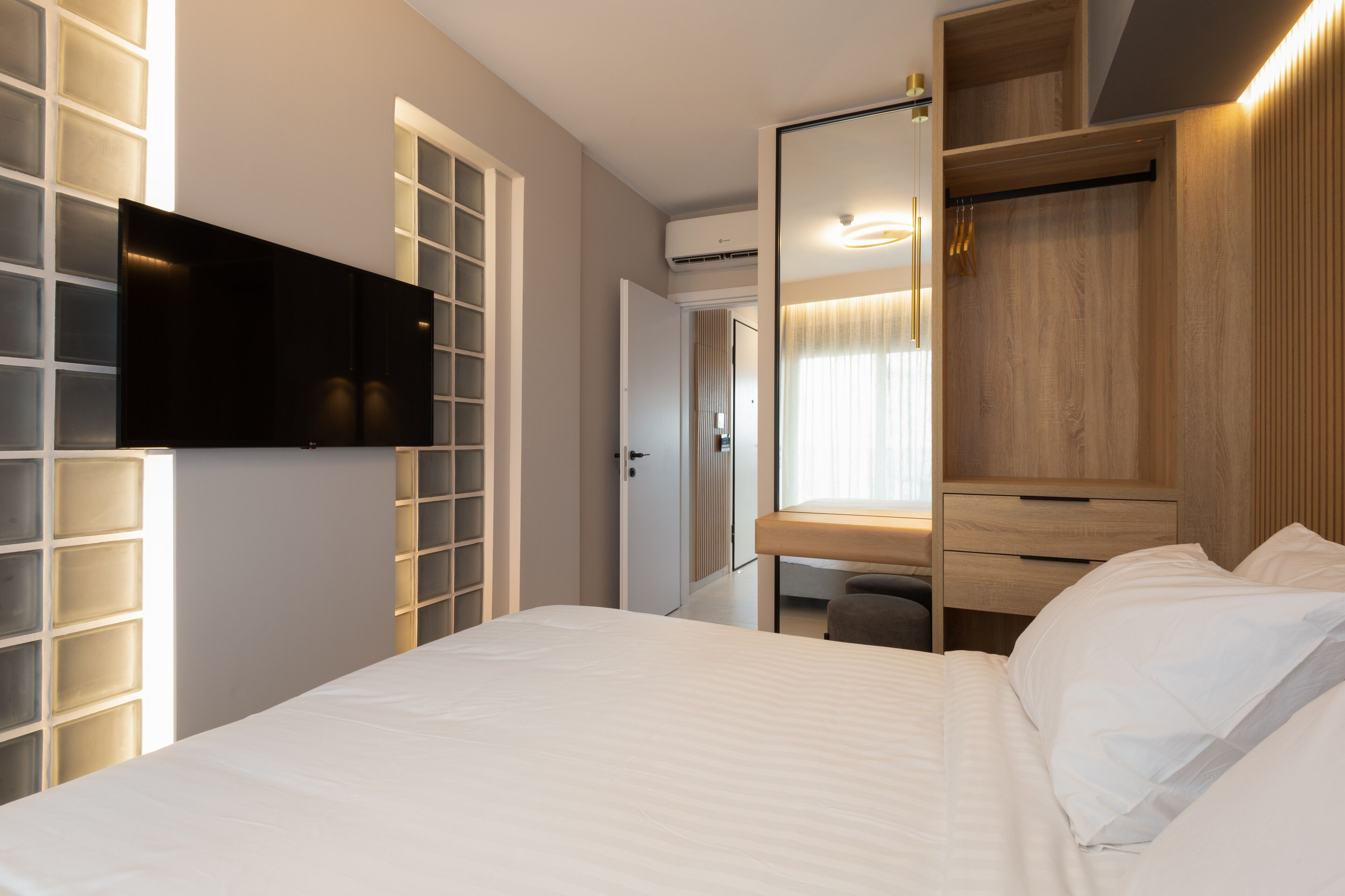 A hotel room with a bed, a TV mounted on the wall, a wooden wardrobe, and a desk with a chair.