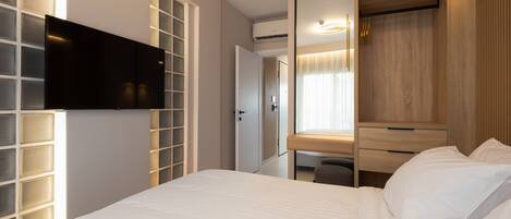 A hotel room with a bed, a TV mounted on the wall, a wooden wardrobe, and a desk with a chair.