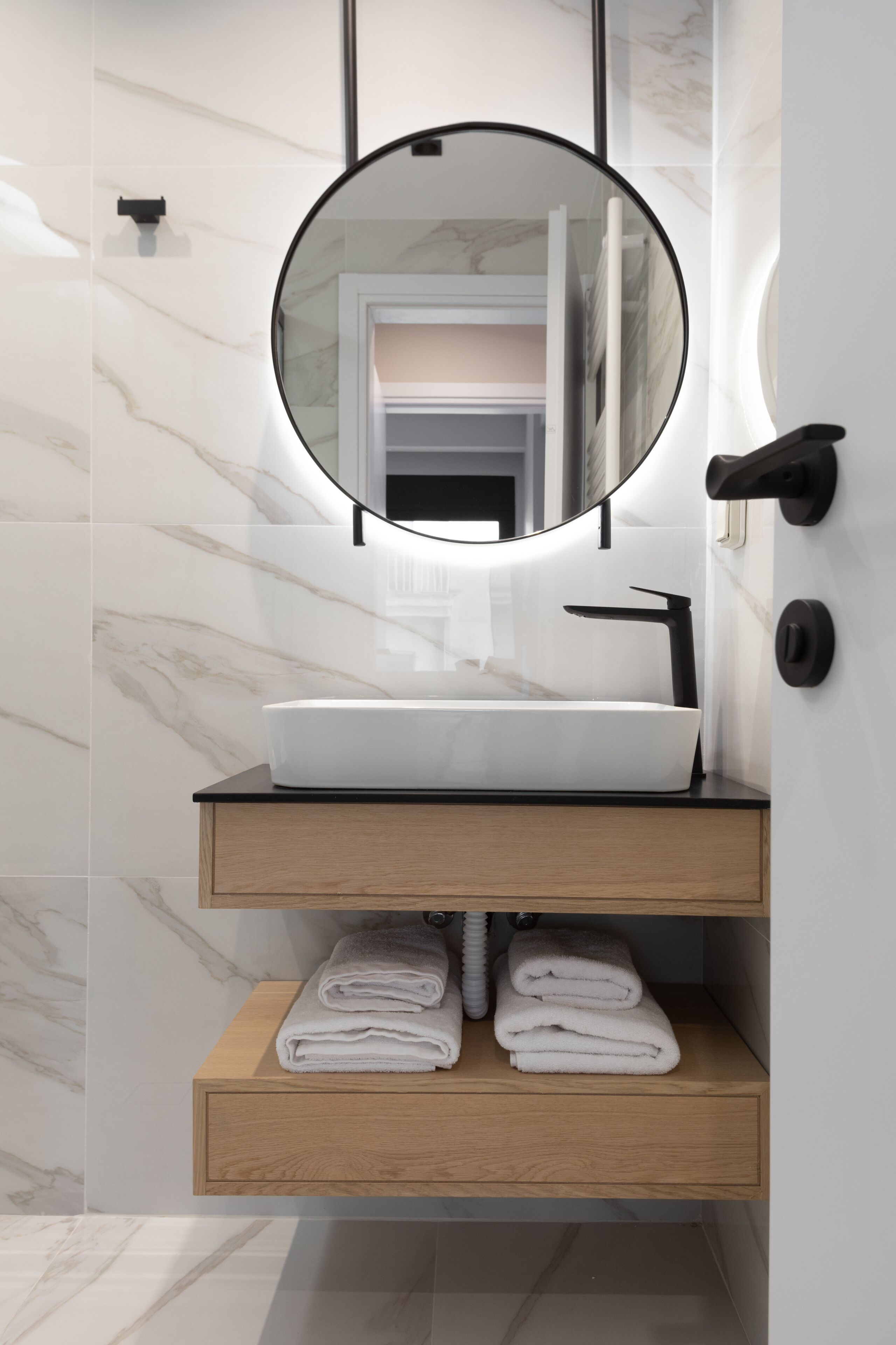 Une salle de bain moderne avec un pare-feu en marbre blanc, un miroir rond, un lavabo blanc et des étagères en bois où sont pliées des serviettes.