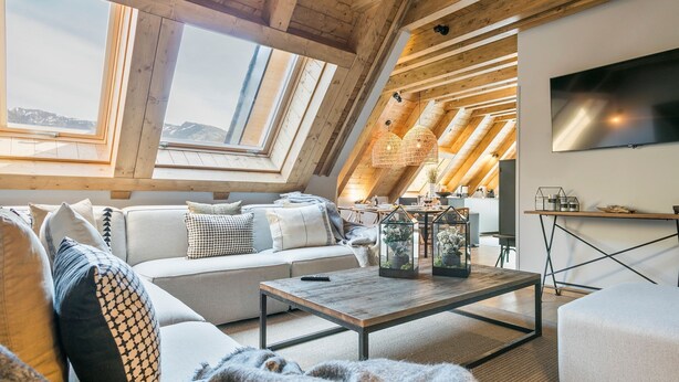A cozy loft-style living room with a high ceiling, wooden beams, a sofa set, a coffee table, and a TV mounted on the wall. - Luderna - Casa Peregrí (Arties)