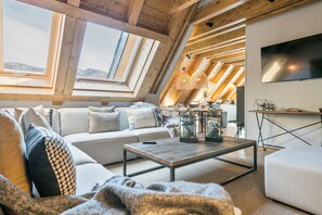 A cozy loft-style living room with a high ceiling, wooden beams, a sofa set, a coffee table, and a TV mounted on the wall. - Luderna - Casa Peregrí (Arties)