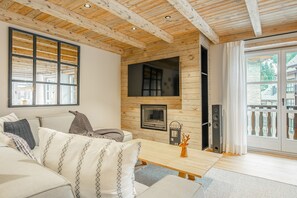 A cozy living room with a wooden ceiling, a fireplace, a sofa with patterned cushions, and a view of the outside.
